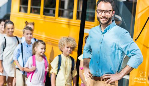Młody mężczyzna na tle żółtego autobusu szkolnego oraz wsiadających do niego dzieci.