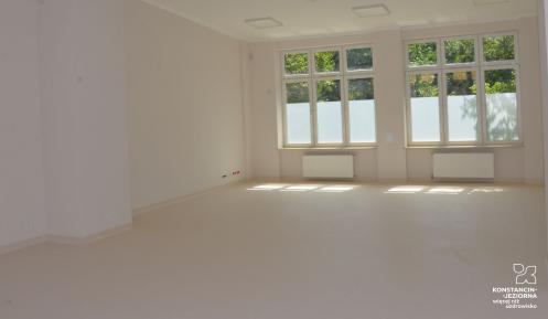 Zdjęcie pomieszczenia, sali pomalowanej na biało. W dali widać duże przeszklone okna. 