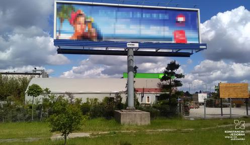  Duży słup reklamowy, na którym umieszona jest kolorowa reklamowa. W tle widać drzewa i budynki. 