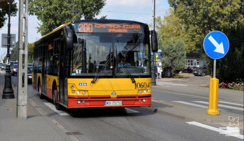 Ulica, po której jedzie czerwono-pomarańczowy autobus komunikacji miejskiej. Nad jego przednią szybą wyświetla się napis: 200 Konstancin-Jeziorna.