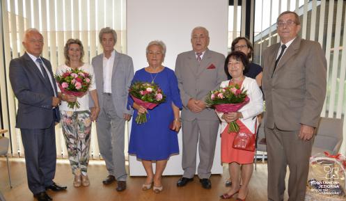 Osiem elegancko ubranych osób stojących frontem do widza, trzy kobiety trzymają kwiaty. Z prawej strony na podłodze stroi kosz prezentowy. Zdjęcie ilustruje sytuacje opisana w artykule.