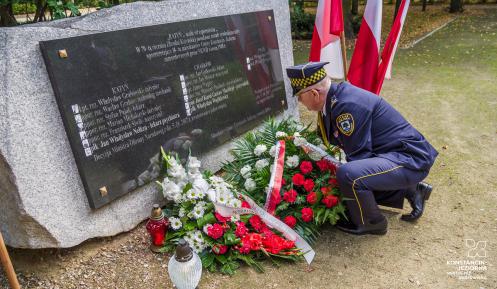  Park. Pomnik ofiar katyńskich. Strażnik miejski składa przed nim wiązankę z białych kwiatów. Obok stoją znicze i trzy biało-czerwone flagi na stojaku. W tle zielone drzewa.