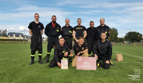 Zdjęcie grupowe 9 strażaków, ubranych w czarne mundury. Stoją w 2 rzędach. Jeden z nich trzyma w rękach duży czek o wartości 1 tys. zł. 