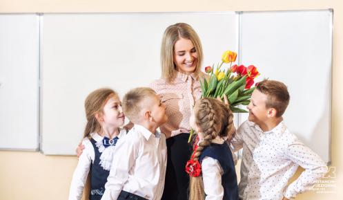 Sala lekcyjna. Czterech uczniów w wieku 10 lat składa życzenia i bukiet swojemu nauczycielowi.