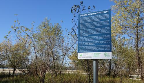 Niebieska tablica a niej białą czcionką podane są liczne zasady obowiązujące na terenie rezerwatu. W tle widać drzewa i rzekę.  