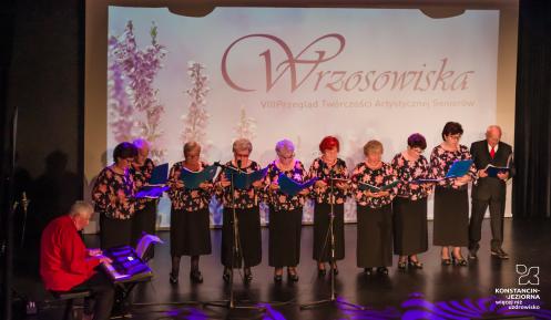 Na scenie występuje seniorzy. 9 kobiet i 1 mężczyzna trzymają w rękach śpiewniki.  W tle wyświetla się napis Wrzosowiska.