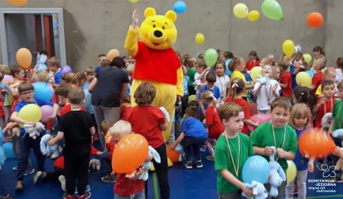 Pluszowa maskotka w towarzystwie dzieci bawiących się balonami.
