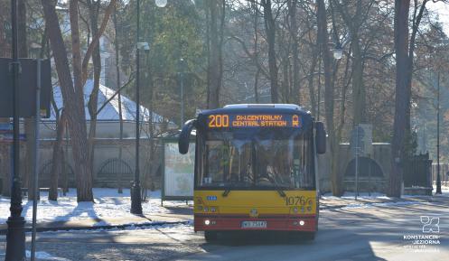 Ulica, po której jedzie czerwono-pomarańczowy autobus komunikacji miejskiej. Nad jego przednią szybą wyświetla się napis: 200. 