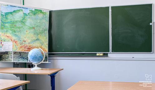 Wnętrze klasy. Na ścianie tablica, przed którą stoją szkolne ławki i krzesła. Na biurko stoi globus. 