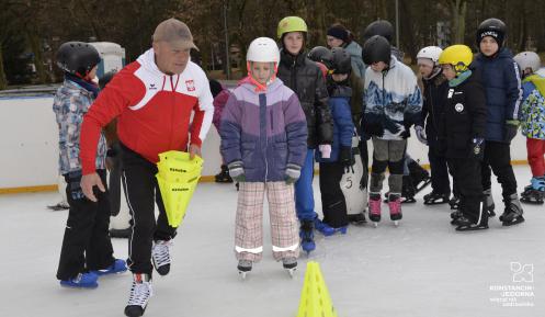Na zdjęciu na lodowisku:  w pierwszym planie mężczyzna na łyżwach w biało-czerwonej kurtce; za nim stoją dzieci też na łyżwach, na lodzie, w kaskach. jedno za drugim.