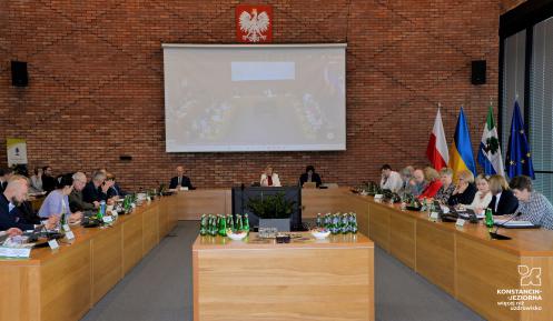 Posiedzenie członków Rady Miejskiej oraz władz gminy. Osoby siedzą przy stołach ułożonych na kształt podkowy. Na środku sali stoi duży telewizor, a na ścianie wisi biały ekran. 