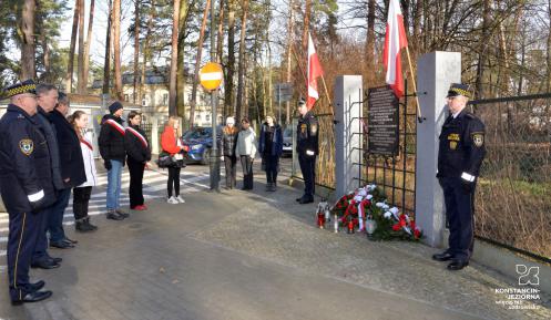 Grupa kilkunastu osób stoi przed pomnikiem pamięci, umieszczonym na ogrodzeniu Białego Dworku. Składają kwiaty i znicze. 