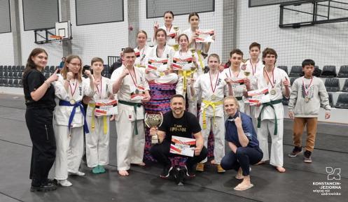 Młode dziewczyny i chłopcy w strojack zawodników karate. Na szyjach mają zawieszone medale, a w dłoniach dyplomy. Przed nimi kuca męzczyzna z pucharem w dłoni i kobieta.