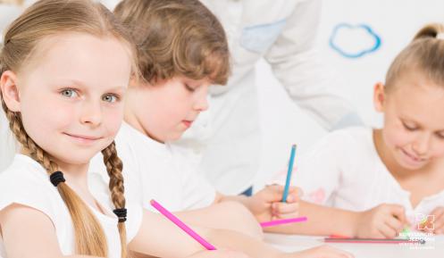 W pierwszym planie dziewczynka z warkoczykami lekko się uśmiecha, w tle chłopiec i dziewczynka spoglądają w zeszyt i coś piszą. W dłoniach mają kolorowe ołówki.