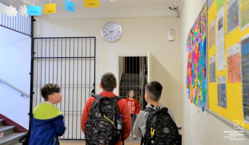Szkolny korytarz, trzech uczniów patrzy w górę na zegar zawieszony na ścianie. 