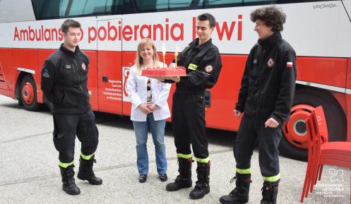 Trzech młodych mężczyzn w kombinezonach strażackich. Jeden z nich trzyma tort. Między nimi młoda kobieta w białym fartuchu.