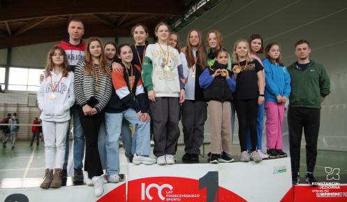 Na podium stoi 13 dziewcząt z medalamizawieszonymi na szyi.  Po prawej i lewej stronie grupy, dwóch mężczyzn w sportowych dresach.