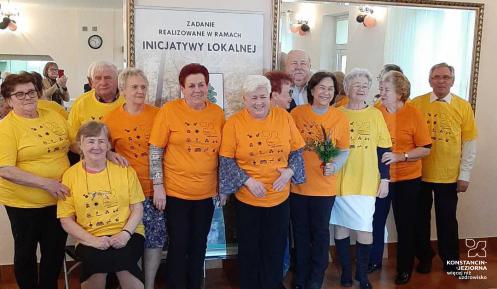 Grupa dwunastu starszych osówb, kobiet i męzczyzn. Sa w żółtych i pomarańczowych koszulkach. Za nimi jest plakat z napisem: zadanie realizowane w ramach inicjatywy lokalnej.