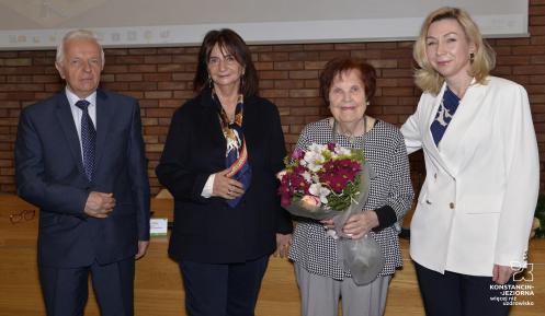 Na zdjęciu stoją trzy kobiety (jedna starsza trzyma w dłoniach bukiet kwiatów). PO lewej stronie stoi starszy mężczyzna, za nimi ceglana ściana i zawieszony ekranem.