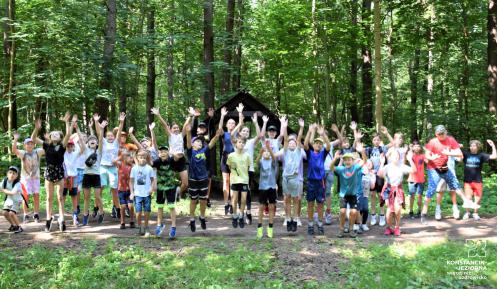 30 dzieci w lesie skacze do góry z wyciągniętymi rękami. 