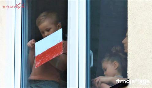 Dziecko stoi w oknie i trzyma w ręku biało-czerwoną flagę. 