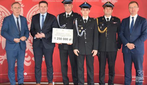 Sześciu mężczyzna, w tym trzech w mundurach strażackich, stoi na tle czerwonej ścianki promocyjnej Państwowej Straży Pożarnej. Jedna osoba trzyma prostokątną tabliczkę, na której widnieje kwota 1 250 000 zł.