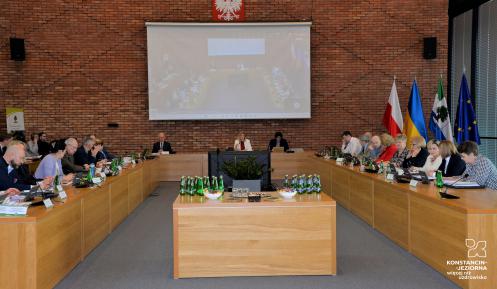 Posiedzenie członków Rady Miejskiej oraz władz gminy. Osoby siedzą przy stołach ułożonych na kształt podkowy. Na środku sali stoi duży telewizor. W tle, na ścianie wyświetlony jest duży ekran.