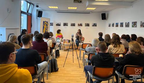 Sala z krzesłami i ludźmi, którzy na nich siedza. Sa tyłem. Przed nimi siedza na krzesłach dwie kobiety, trzymają w dłoniach mikrofony.