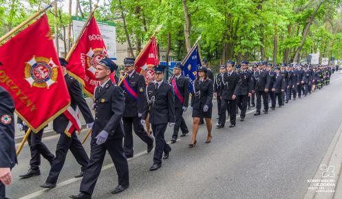 Strażacy maszerują ulicą. Za nimi zielone drzewa. W dłoniach trzymają sztandary.