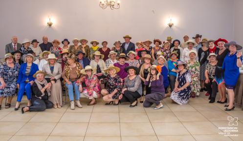 Ponad 60 starszych osób (kobiety i mężczyźni) w dużej sali, ustawionych do zdjęcia. Wszyscy sa w kapeluszach i kolorowo ubrani.