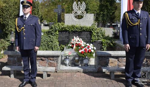 Dwóch mężczyzn w mundurach strazy miejskiej stoi czed grobem, który jest ogrodzeny niskim murkiem. Na tablicy wypisane są imiona i nazwiska oraz  napis „1939, poległym za ojczyznę”. 