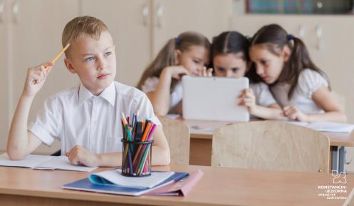W sali za szkolnym stołem siedzi chłopiec w białej bluzce. Ma ołówek przy głowie. Na stole leżą zeszyty i kredki. Za nim, również za szkolnym stołem, siedzą trzy dziewczynki i patrzą w monitor komputera.