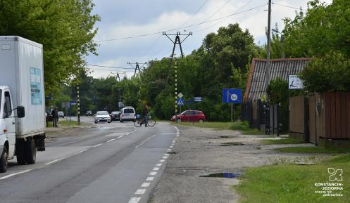 Zniszczona asfaltowa droga bez pobocza. Jedzie po niej ciężarówka, samochody osobowe oraz przechodzi przez nia mężczyzna z rowerem.