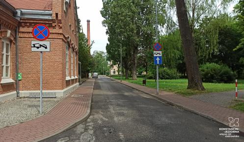 Ulica przy kturej stoi ceglany budyne oraz znak drogowy z zakazem parkowania i mozliwościa odholowania pojazdu.