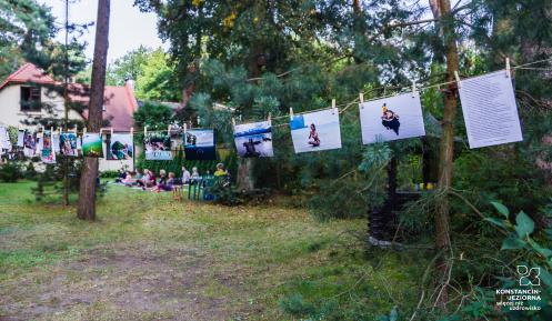 Działka, kilka wysokich drzew, a między nimi rozwieszone na sznurku zdjęcia. W tle widać grupę osób, która siedzi na trawie i uprawia jogę. 