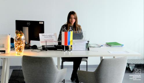 Młoda kobieta siedzi za dużym stołem, na którym stoją dwie flagi - polska i ukraińska oraz leżą dokumenty,