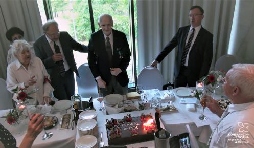 Za stołem stoi starszy mężczyzna, a obok niego stoją trzy osoby. Na stole stoi tort ze świeczkami.