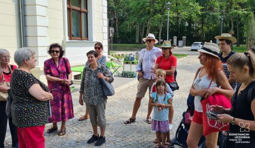 Przestrzeń publiczna. Grupa osób stoi przed budynkiem, patrzy na przewodnika.