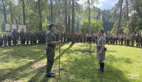 Na leśnej polanie stoją naprzeciwko siebie mężczyzna z kijem w ręce, którym się podpiera oraz dziewczyna. Są w harcerskich mundurach. W oddali na baczność stoi kilkadziesiąt osób  –  dziewczyny i chłopcy również w harcerskich mundurach.