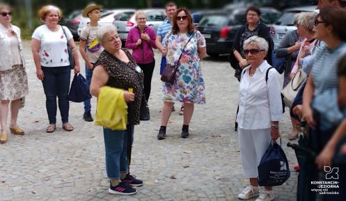 Przestrzeń publiczna. Grupa osób stoi przed budynkiem, patrzy na przewodnika.