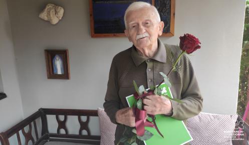 Staruszek trzyma w ręku dużą czerwoną róże i zieloną teczkę