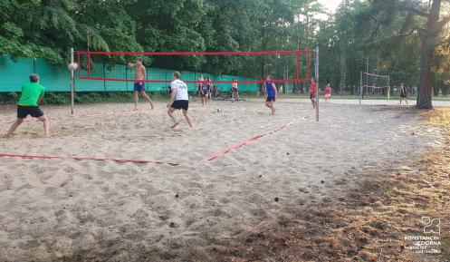 Na piasku grupa mężczyzn odbija piłkę. Dwóch znajduje się po jednej stronie siatki, a dwóch po drugiej. Są boso i w strojach sportowych..