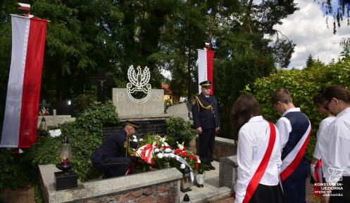 Dwóch strażników miejskich pełni wartę przy nagrobku na cmentarzu. Przed nageobkiem młodzi ludziewbiało-czarnych strojach. 