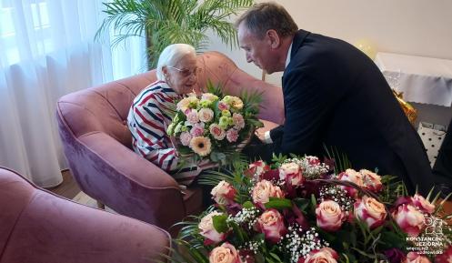 Starsza kobieta siedzi na sofie. Pochyla sie do niej mężczyzna w garniturze. Ma w dłoni bukiet kwiatów.