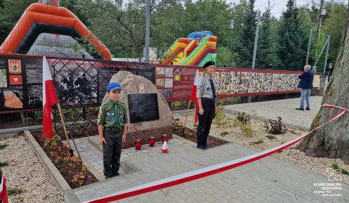 Chłopiec i dziewczynka, ubrani w mundury harcerskie, stoją na baczność przy głazie z białym orłem i czarna tablicą. Za nimi ogrodzenie, na którym wiszą zdjęcia, a dalej dmuchańce.