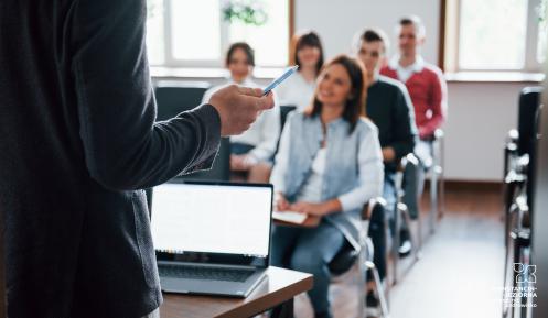 Grupa ludzi w nowoczesnej klasie, siedzą na krzesłach za stolikami. Wszyscy się uśmiechają i słuchają. Na pierwszym planie widać sylwetkę mężczyzny. 