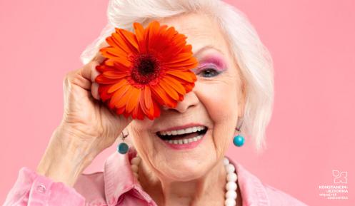 Usmiechnieta seniorka trzymająca zcerwony kwiatek na wysokości oka na różowym tle.
