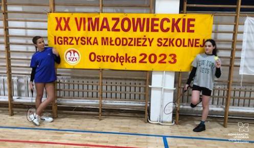 Dwie uczennice spozuja na tle baneru informującym o 26. Mazowieckich Igrzyskach Młodzieży Szkolnej.