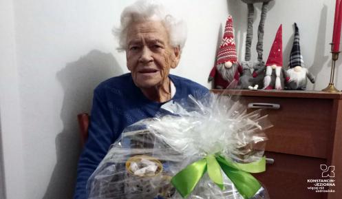 Starsza kobieta trzyma kosz z produktami spożywczymi, zapakowany w folię dekoracyjną.