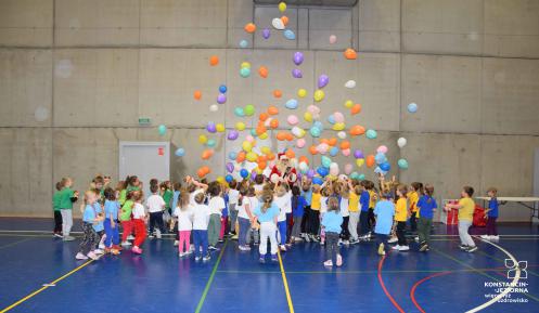 Przedszkolaki stoją na hali sportowej zebrane do okoła Mikołaja. Nad nimi lataja balony. 
